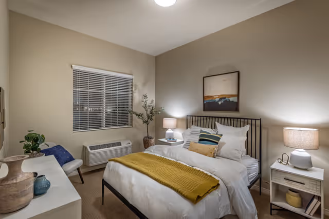 Well-lit furnished bedroom with a bed, nightstands and lamps, a dresser, window and decorative plants.