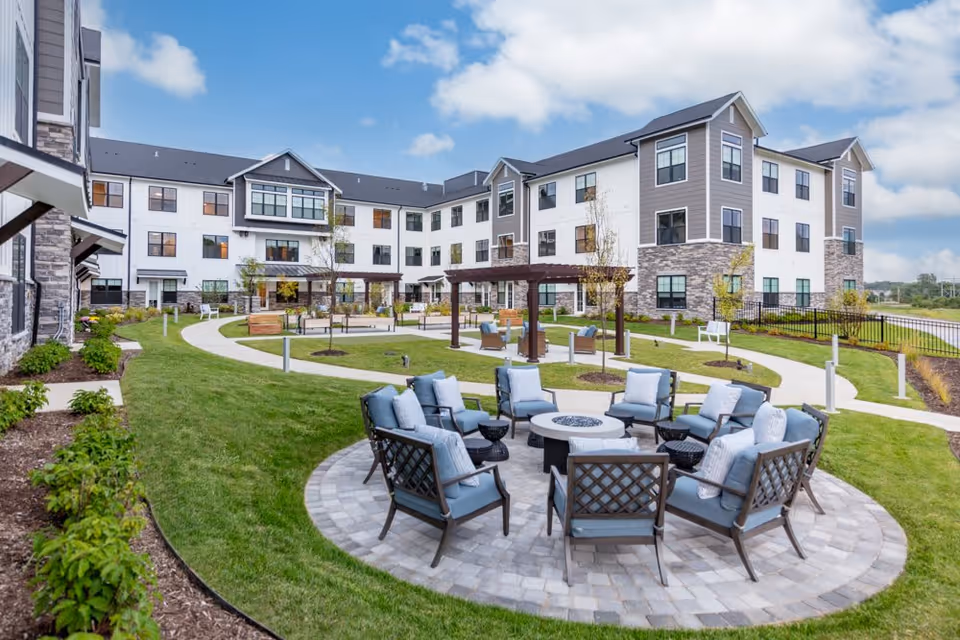 Outdoor courtyard area of a senior living facility with a circular seating arrangement of cushioned chairs around a fire pit, surrounded by green grass, pathways, small trees, and a three-story building in the background under a partly cloudy sky.