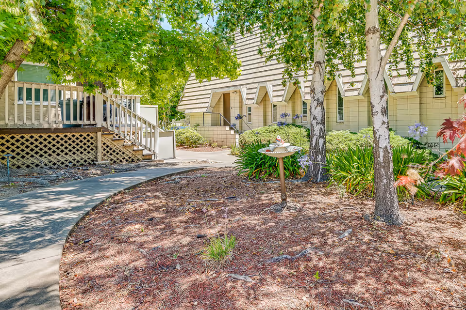 Outdoor garden area at Oakland Heights Senior Living with a curved concrete pathway, trees, green shrubs, and a birdbath in the center. The building exterior with beige siding and triangular roof accents is visible in the background.