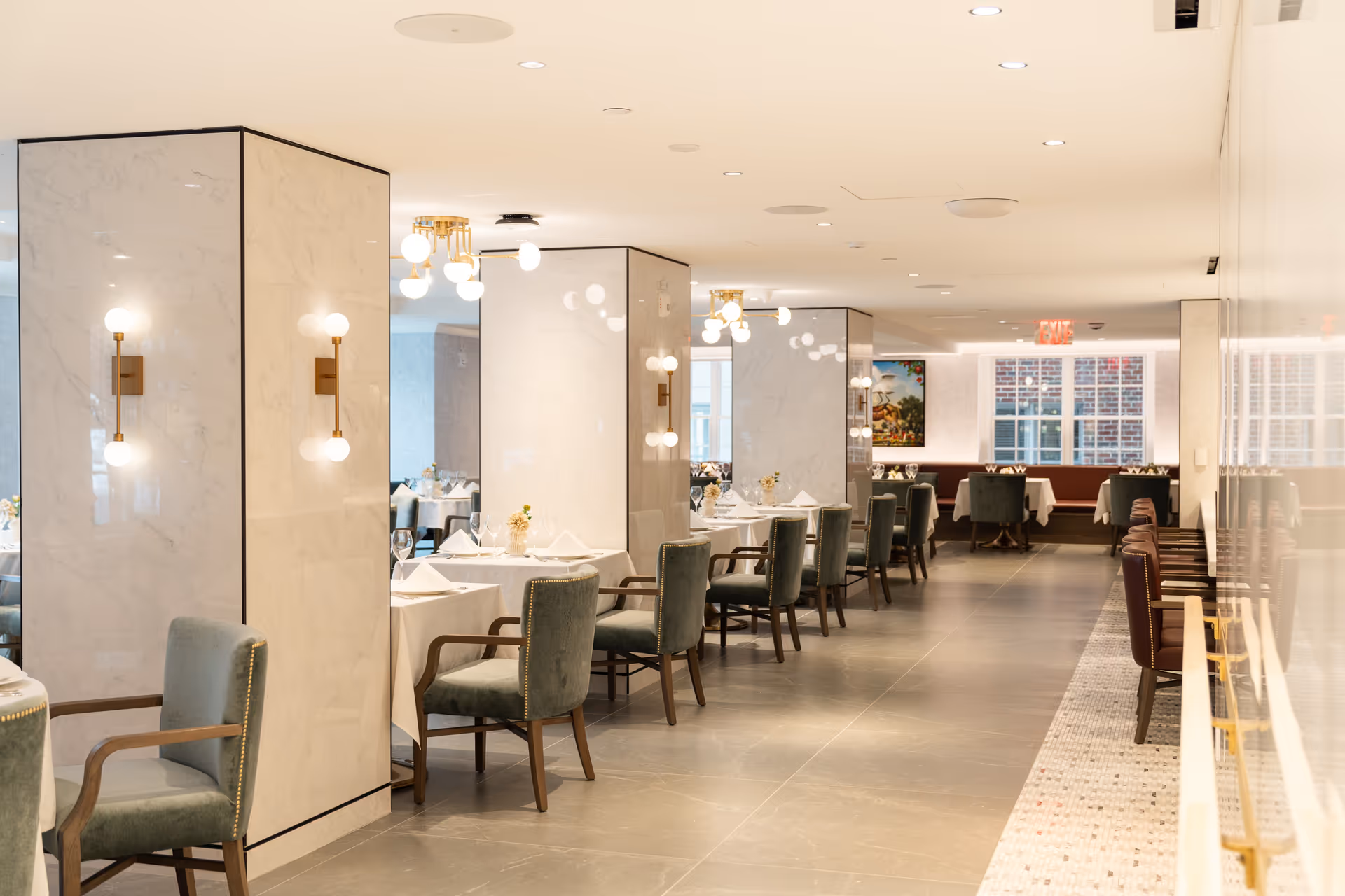 A bright, elegant dining room with tables covered in white tablecloths, set with glassware and napkins. The room features marble pillars with modern wall sconces, green upholstered chairs, and large windows letting in natural light. The floor is tiled, and there are decorative ceiling lights.