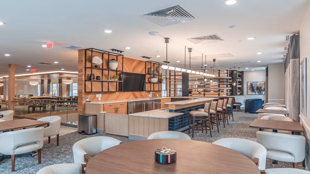 A modern, spacious common area in a senior living facility featuring a large round wooden table with white cushioned chairs around it in the foreground. In the background, there is a long counter with bar stools, a wall-mounted TV, decorative shelves with vases and plants, and additional seating areas with tables and chairs. The room is well-lit with ceiling lights and pendant lighting above the counter.