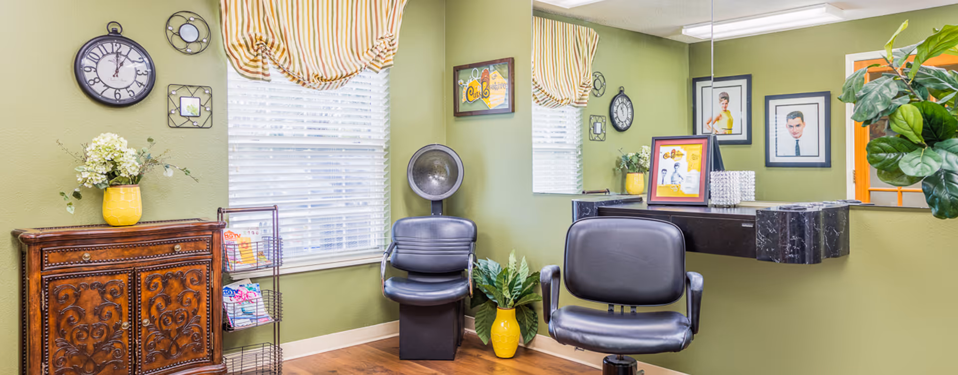 A bright interior salon area with two black styling chairs, a hooded hair dryer, a wall mirror, decorative cabinet and plants against green walls.