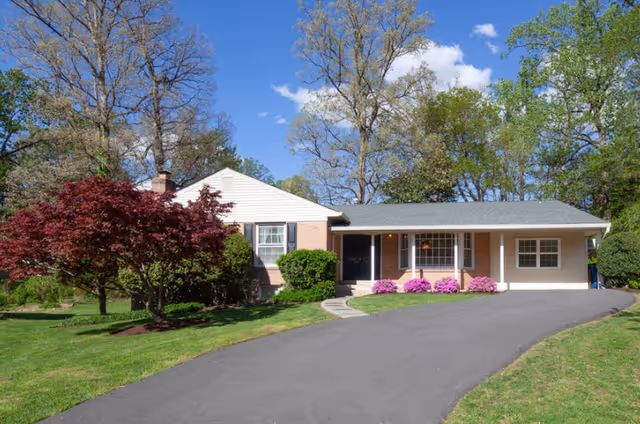 Single-story house with a sloped driveway, surrounded by green grass, trees, and flowering bushes under a blue sky with some clouds.