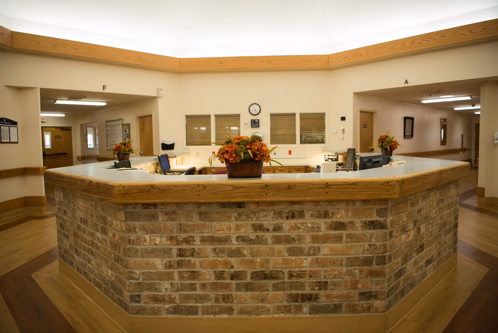 Reception desk area in a senior living facility with a brick front and wooden trim. The desk has computer monitors, office supplies, and flower arrangements. Behind the desk are windows with blinds, a clock, and hallway entrances labeled A and F.