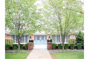 Front exterior view of a senior living facility with a pathway leading to double blue doors, flanked by brick pillars and trimmed hedges, with two large leafy trees on either side.