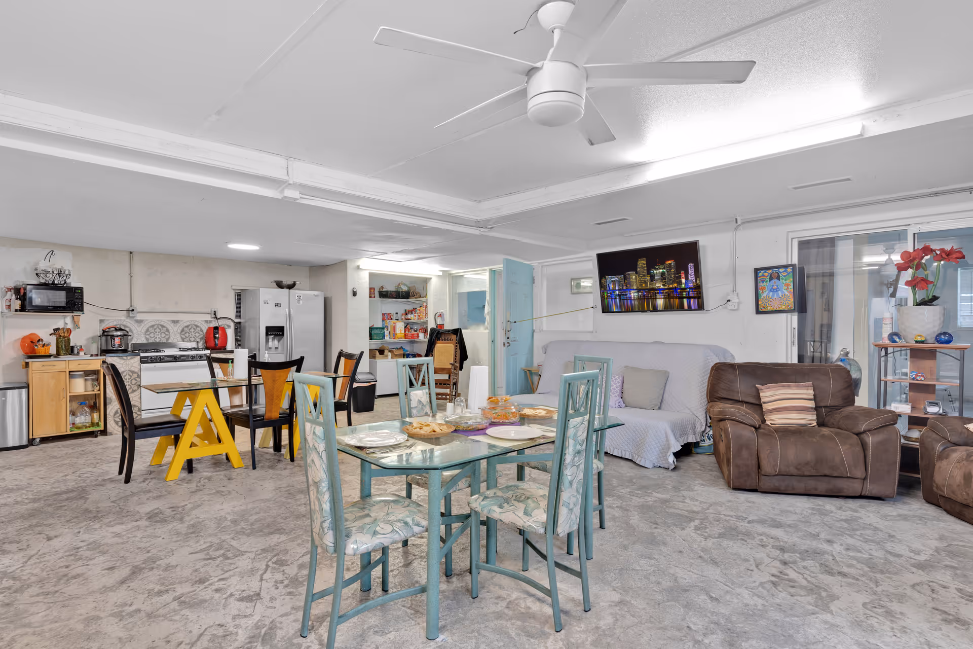 A combined living and dining area with a glass dining table and four chairs in the foreground, a small kitchen area with appliances including a refrigerator, stove, microwave, and kettle in the background, and a seating area with a couch covered with a light-colored slipcover and two brown armchairs. The room has a concrete floor, white walls, a ceiling fan, and a wall-mounted TV displaying a cityscape at night. There are some decorative items and artwork on the walls and shelves.