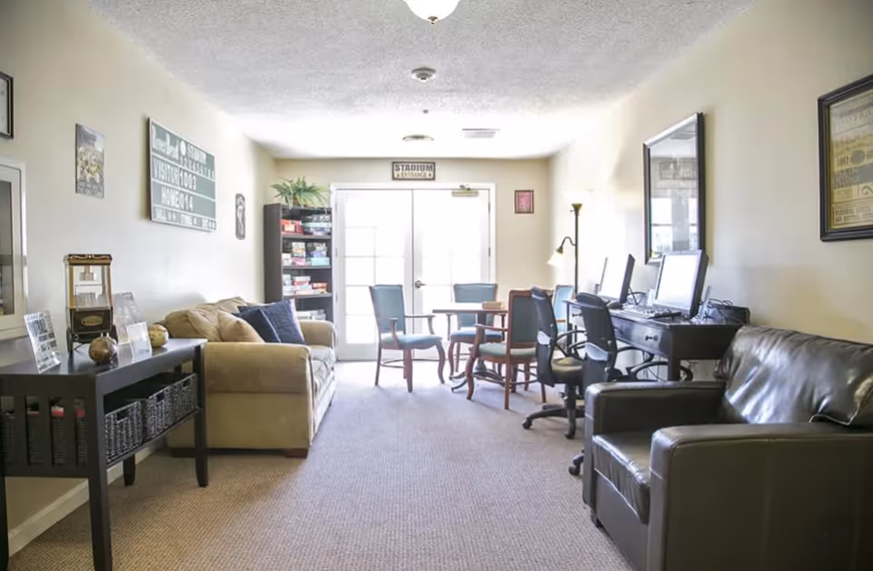 A well-lit common area with a beige sofa and a black leather armchair on opposite sides. There is a black console table with decorative items on the left, a bookshelf filled with board games near the back wall, and a table with four chairs near a set of double glass doors. On the right side, there is a desk with three computer monitors and office chairs. The walls are decorated with framed pictures and signs.