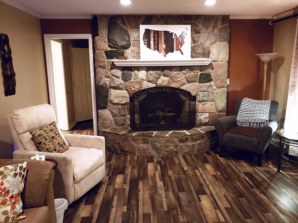 Cozy living room with a stone fireplace in the center, flanked by a beige armchair with a patterned pillow on the left and a gray armchair with a striped throw blanket on the right. The room has wood flooring, a floor lamp, and a side table near the window with sheer curtains.