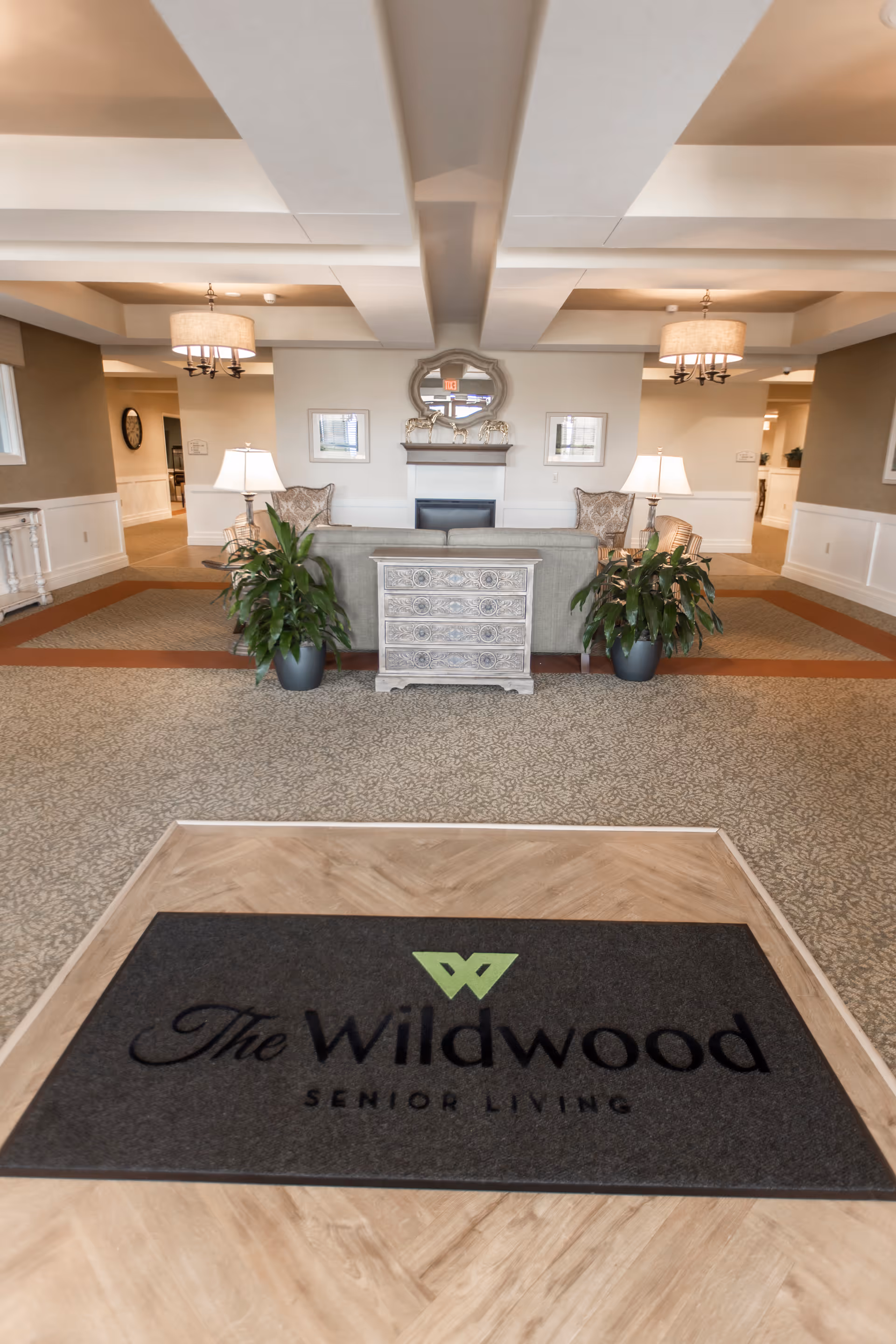 Lobby interior of The Wildwood Senior Living with a branded entrance mat in the foreground, a seating area with sofa and lamps, potted plants, and a mirror above a fireplace.