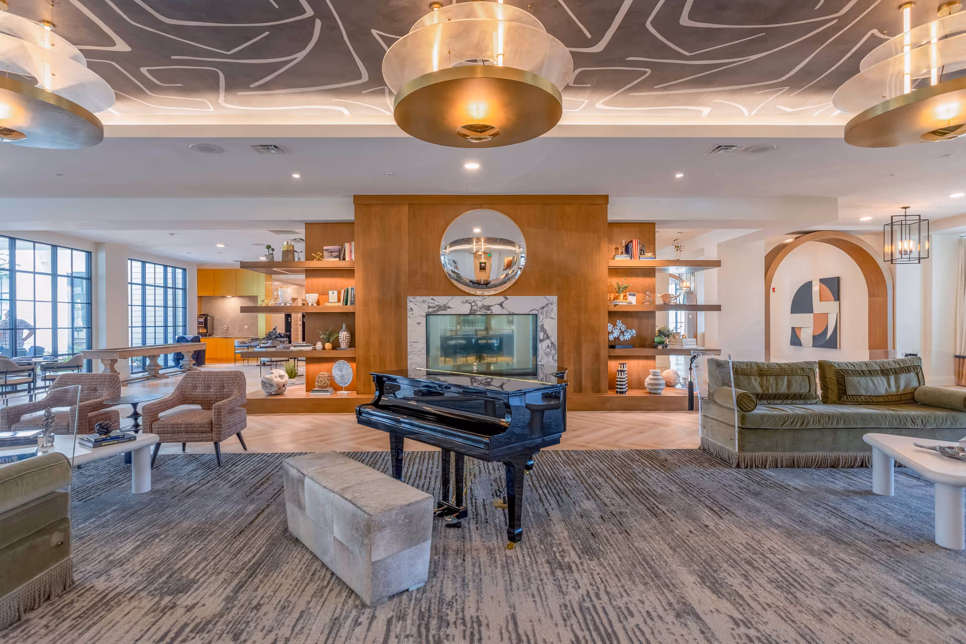 A spacious and modern living room area featuring a black grand piano with a beige cushioned bench in front. The room has a large patterned rug, green velvet sofas, and two beige armchairs around small white tables. The back wall has a wooden panel with a round mirror above a marble fireplace, flanked by open shelves with decorative items. Large windows on the left side allow natural light to fill the space, and the ceiling has artistic lighting fixtures and a unique patterned design.