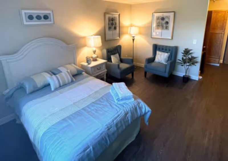 A cozy bedroom with a neatly made bed featuring a light blue and white quilt, two pillows, and folded towels on top. Next to the bed is a white nightstand with a lamp and decorative items. Two blue upholstered armchairs with cushions are placed against the wall, accompanied by a floor lamp and framed artwork above. A potted plant is near the doorway, and the room has wooden flooring and soft lighting.