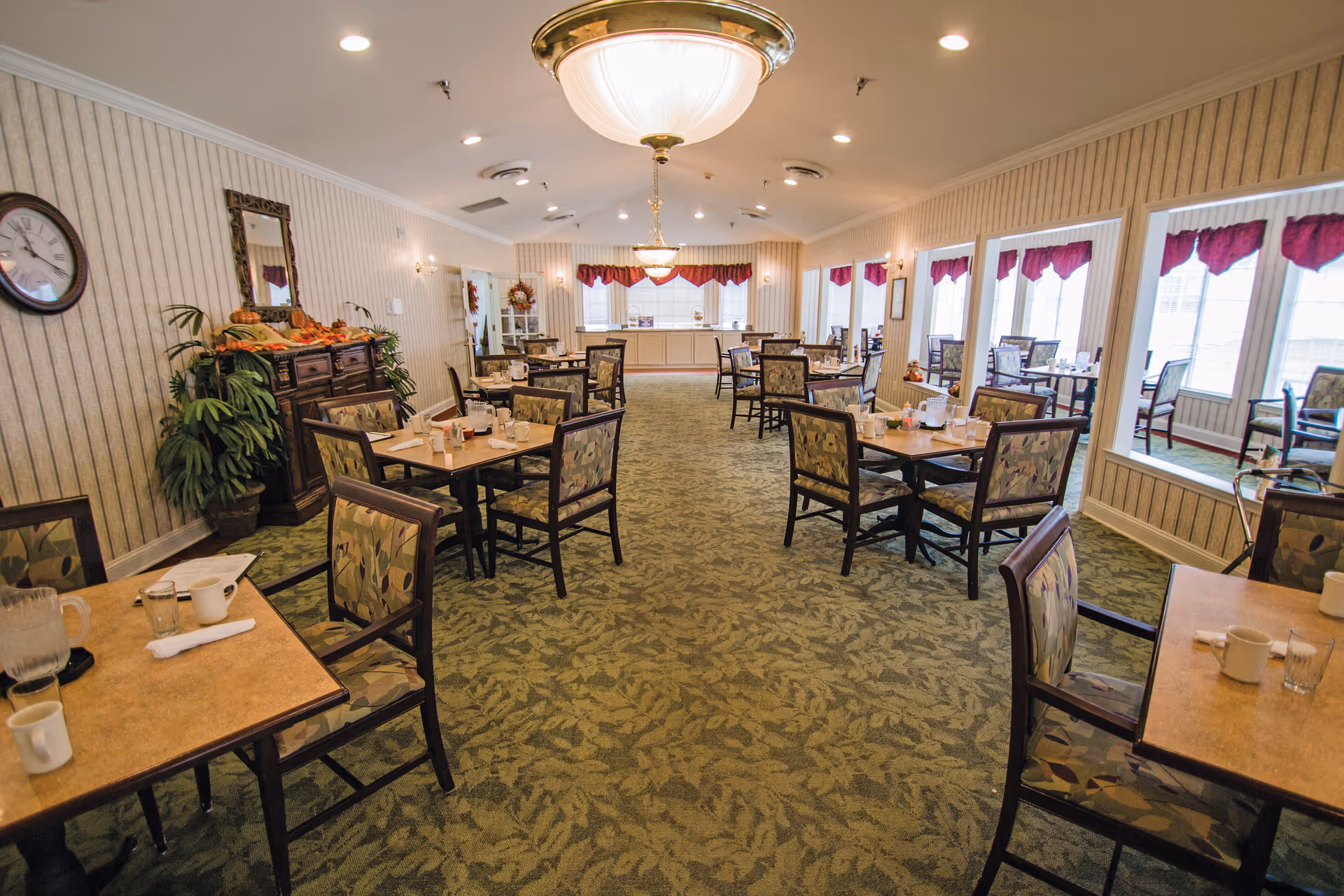 A dining room in a senior living facility with multiple tables and chairs arranged neatly. The room has green patterned carpet, beige striped wallpaper, and large mirrors on one wall. There are decorative plants and a wooden cabinet with autumn-themed decorations. The room is well-lit with ceiling lights and large windows with purple valances.
