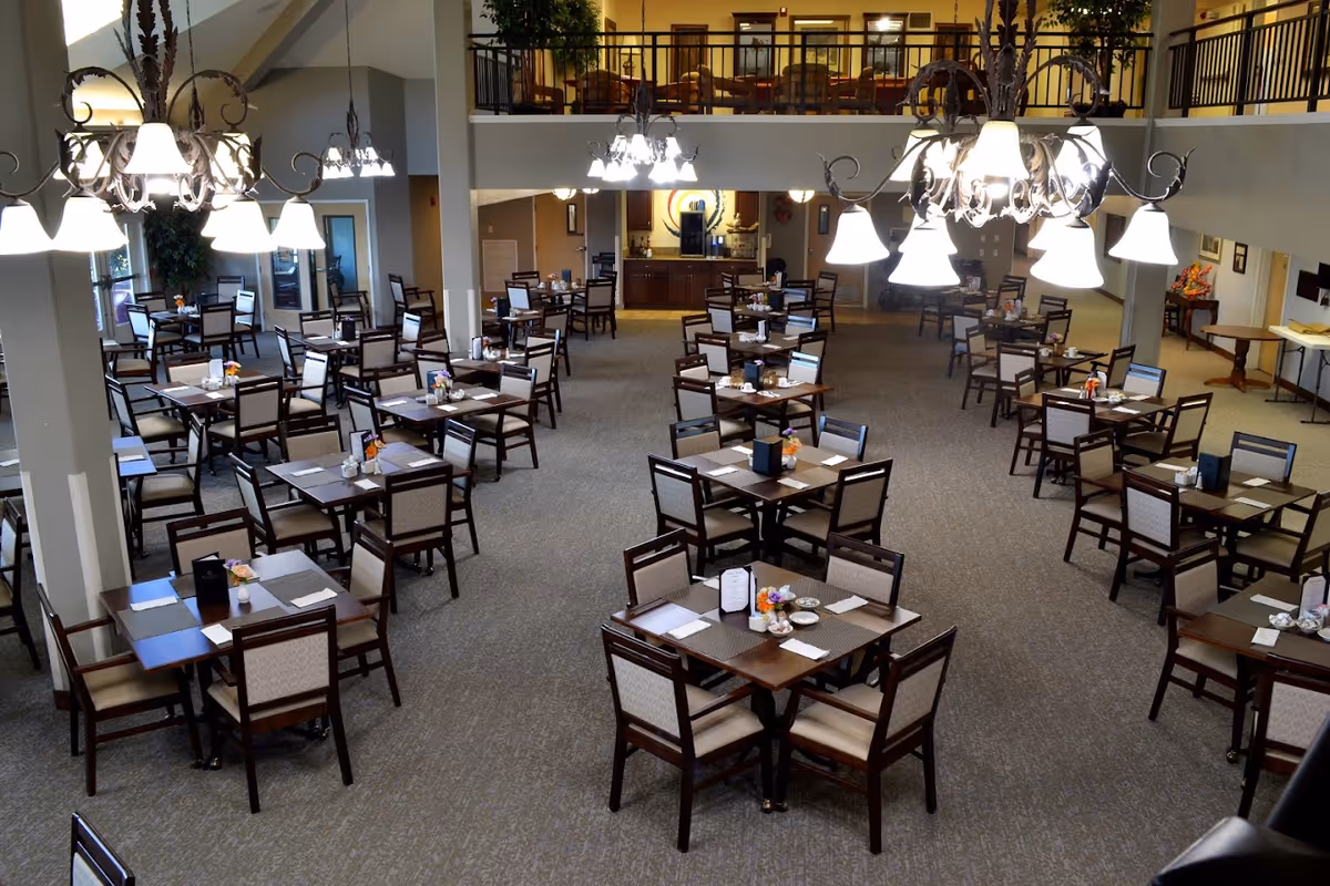 Large dining room with many neatly set tables, chairs, and hanging chandeliers in a senior living facility.