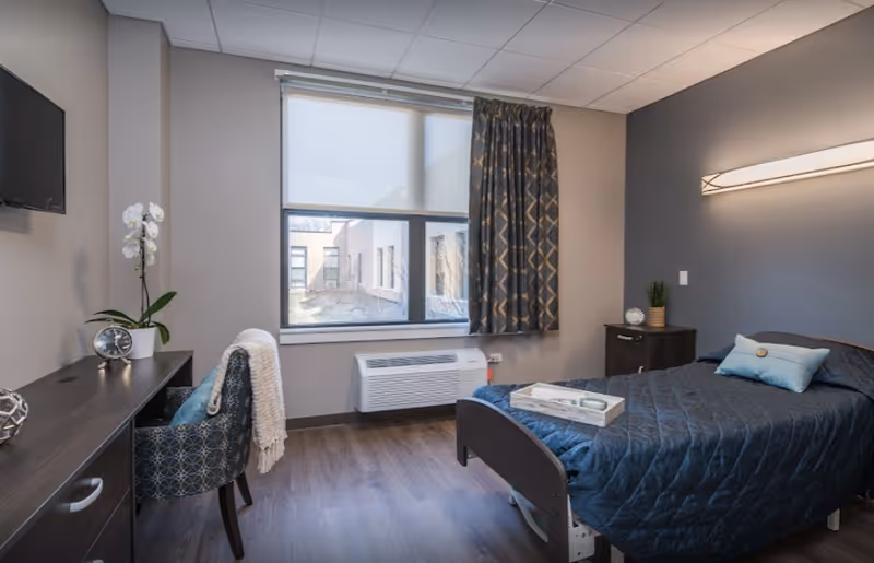 A neatly arranged bedroom in a senior living facility with a single bed covered in a dark quilt and a light blue pillow. There is a small bedside table with a plant and decorative item, a window with patterned curtains and a roller shade, a wall-mounted light fixture, a desk with a chair draped with a white blanket, a clock, and a potted orchid. The room has wood flooring and neutral-colored walls.