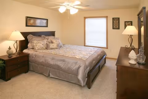 A neatly made bed with a wooden headboard in a bedroom. The bed has a beige and light brown comforter with multiple pillows. There is a wooden nightstand with a lamp and decorative items on the left side of the bed. A window with closed blinds is on the wall behind the bed. On the right side, there is a wooden dresser with a lamp and decorative items. The walls are painted light beige and the floor is carpeted.