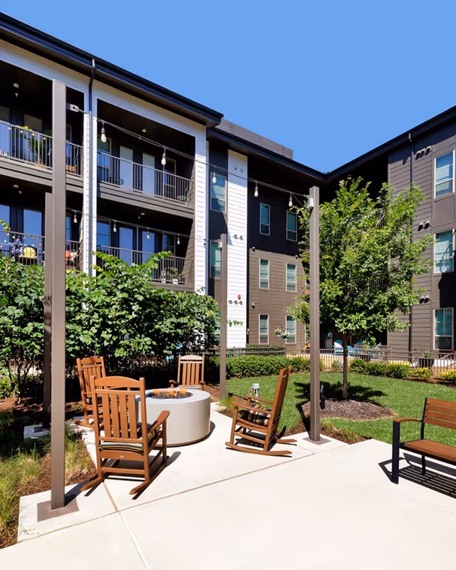 Outdoor courtyard area at Album Benbrook featuring wooden rocking chairs arranged around a circular fire pit, string lights hanging overhead, green bushes, a tree, and a multi-story residential building in the background under a clear blue sky.