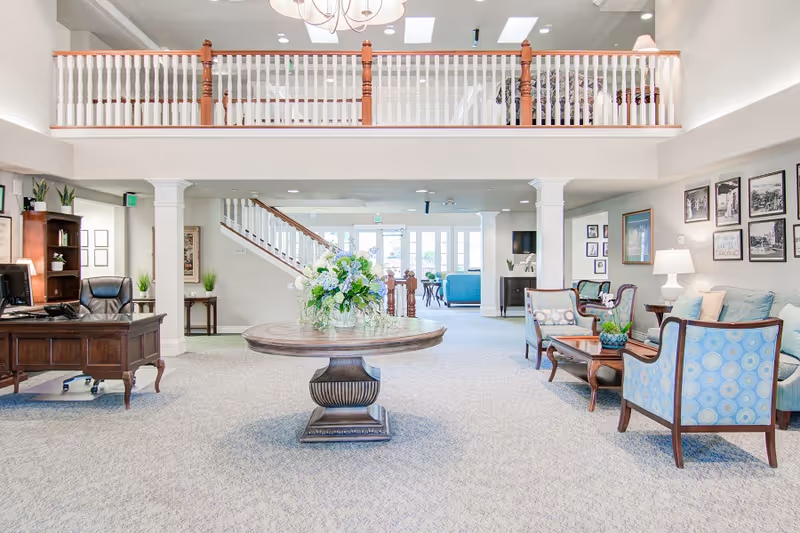 Bright and spacious senior living facility common area with a round wooden table holding a floral arrangement in the center. To the right, there are blue upholstered chairs and a sofa with a wooden coffee table, lamps, and framed pictures on the wall. To the left, there is a wooden desk with a computer and office chair. A staircase with wooden railing leads to an upper balcony overlooking the area.