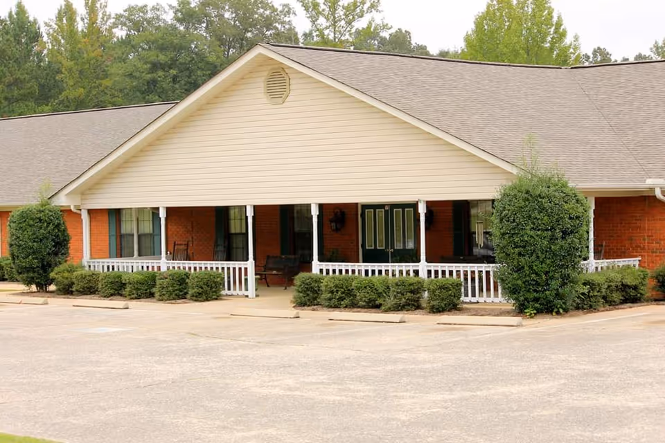 Exterior view of a single-story brick building with a beige gabled roof and a covered porch featuring white railings. The porch has seating including a bench and rocking chairs. There are bushes and small trees planted along the front of the building, and a paved parking area in front. Trees are visible in the background.