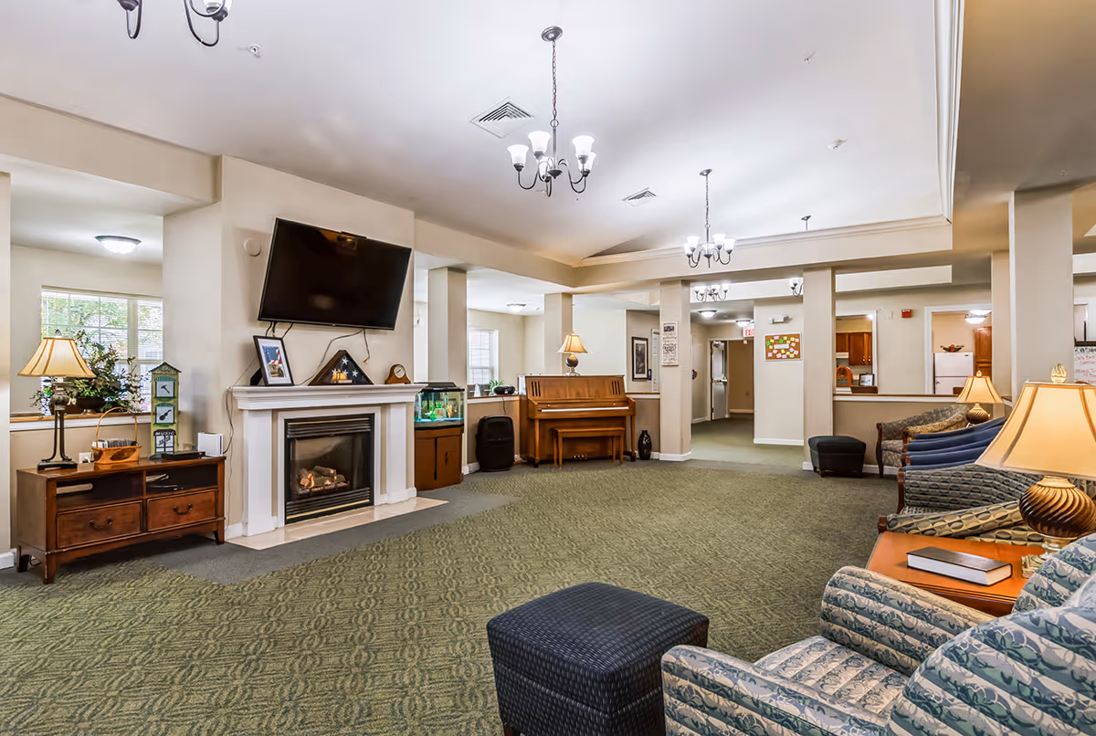 Spacious senior living lounge with a fireplace and mounted TV, piano, seating areas, lamps, and carpeted floor.