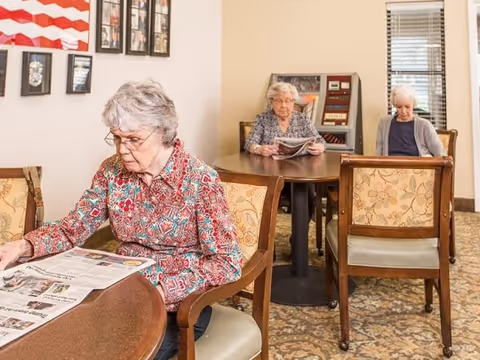 Three elderly women sitting at separate tables in a common area of a retirement community. Two of the women are reading newspapers, and the room has patterned carpet, beige walls, and windows with blinds.
