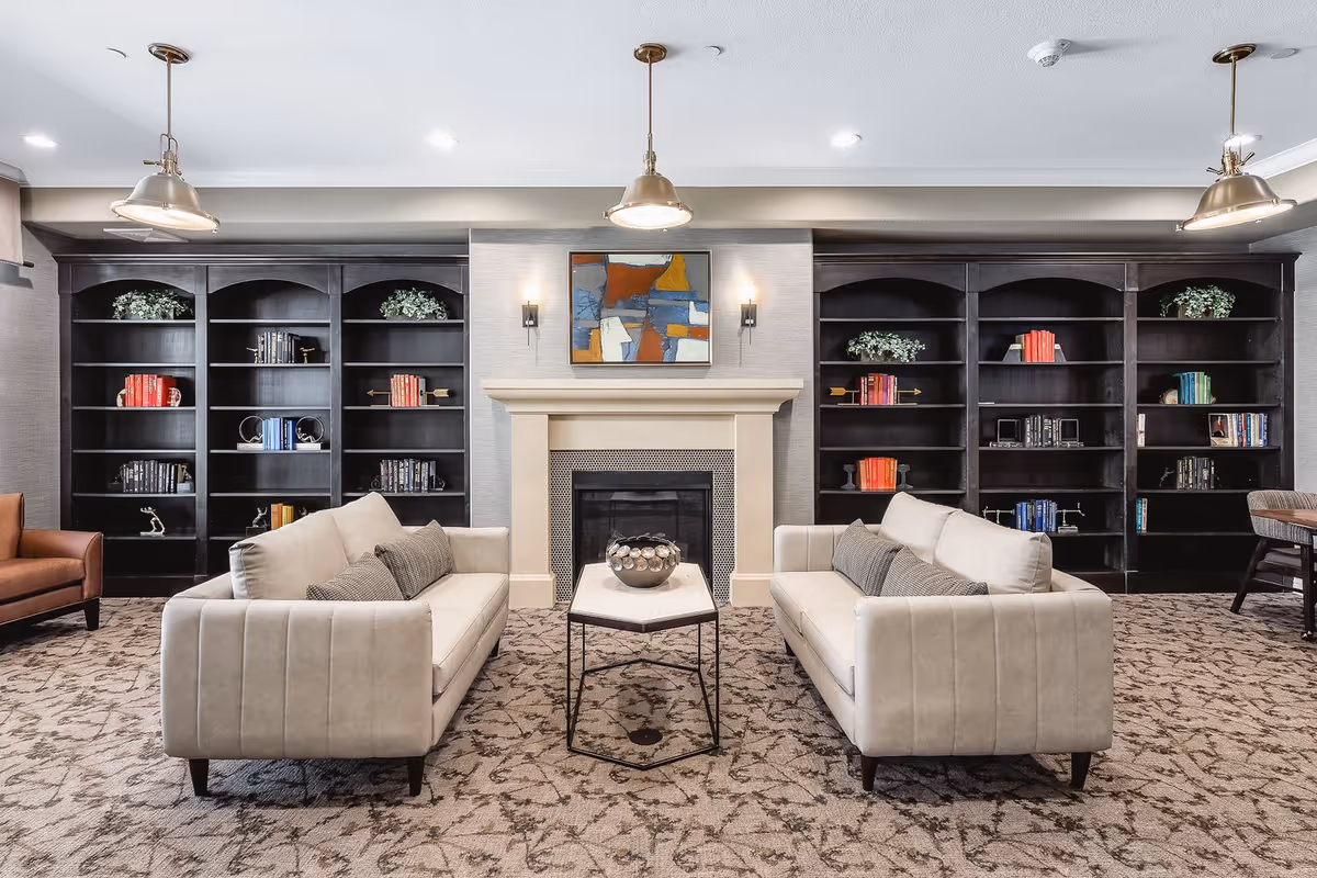 A cozy senior living common area featuring two beige sofas facing each other with a small hexagonal coffee table in between. Behind the sofas is a fireplace with a modern abstract painting above it. On either side of the fireplace are large dark wood bookshelves filled with books and decorative plants. The room is softly lit by three pendant lights hanging from the ceiling and two wall sconces flanking the fireplace. The floor is carpeted with a patterned design.