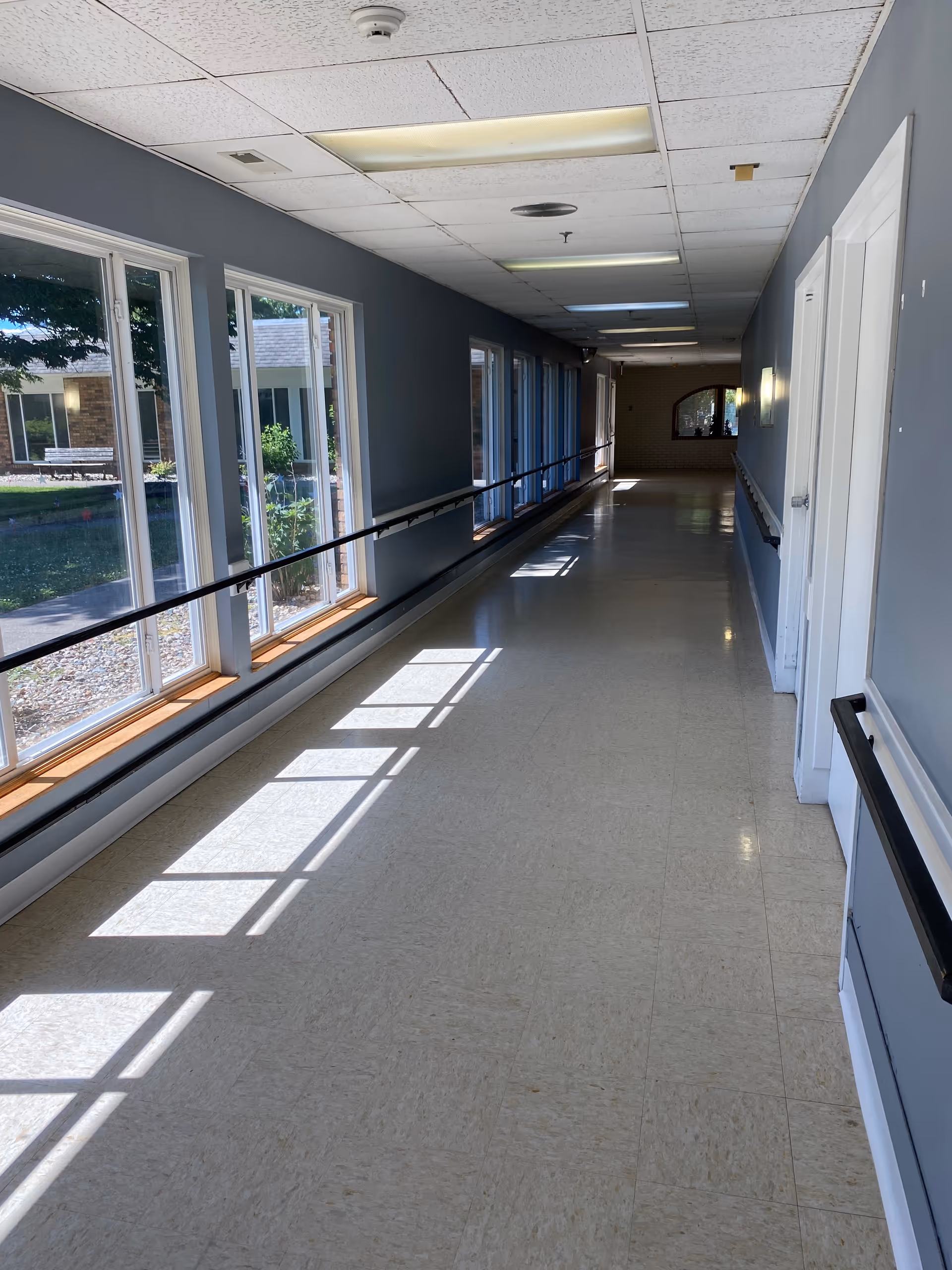 Sunlit long interior hallway with large windows on the left, handrails, and a tiled floor.