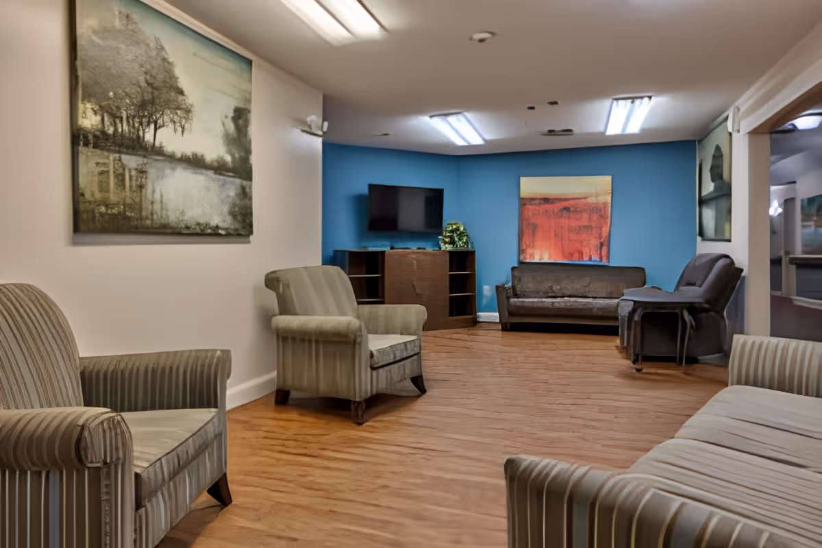A senior living facility common area with wooden flooring, several upholstered armchairs and sofas arranged around the room. There is a blue accent wall with a flat-screen TV mounted on it and a colorful abstract painting. The walls are decorated with framed artwork, and the room is lit by ceiling fluorescent lights.