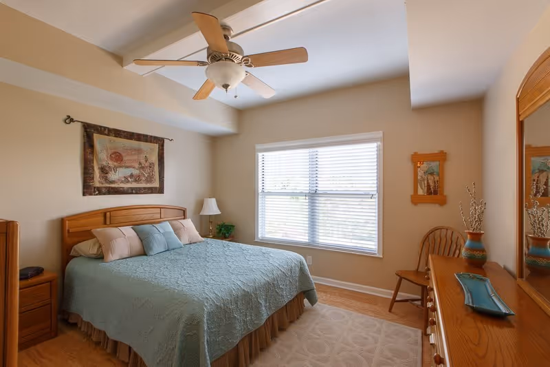A cozy bedroom with a large bed covered in a light blue quilt and several pillows. The room features a wooden headboard, matching wooden nightstand with a lamp, a wooden dresser with a mirror, a wooden chair, and a window with white blinds letting in natural light. A ceiling fan with wooden blades is mounted on the ceiling, and a decorative wall hanging is above the bed.