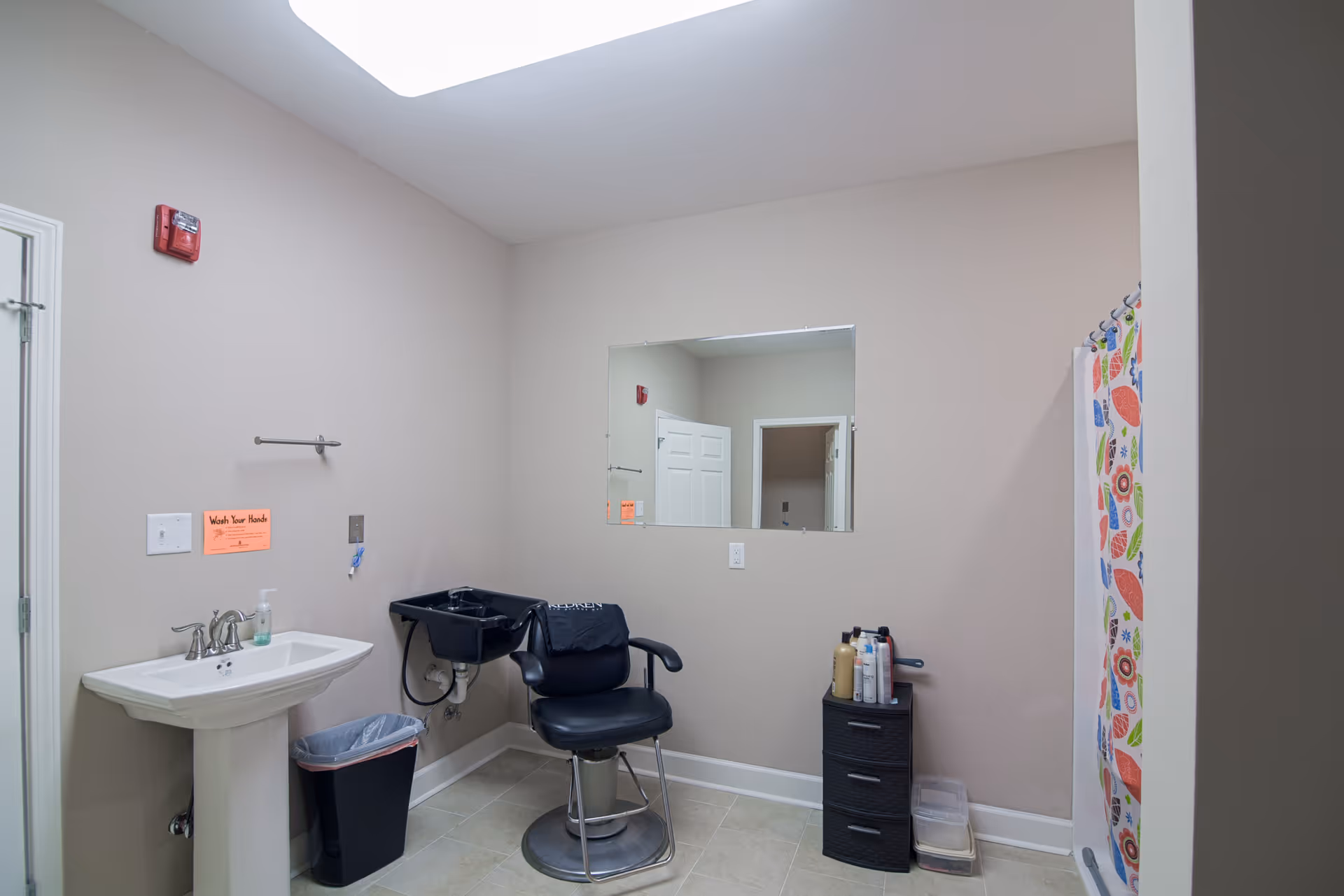 Interior room with a pedestal sink, a black salon chair in front of a black wash basin, a small black drawer unit with hair care products on top, a colorful shower curtain on the right, and a large mirror on the wall reflecting a door.