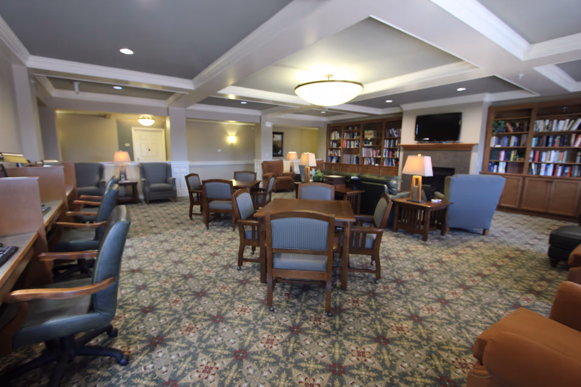 A spacious common area in a senior living facility featuring multiple wooden tables with chairs, several armchairs, and sofas arranged around a fireplace with a mounted TV above it. The room has built-in bookshelves filled with books, a patterned carpet, and a ceiling with recessed lighting and a central light fixture.
