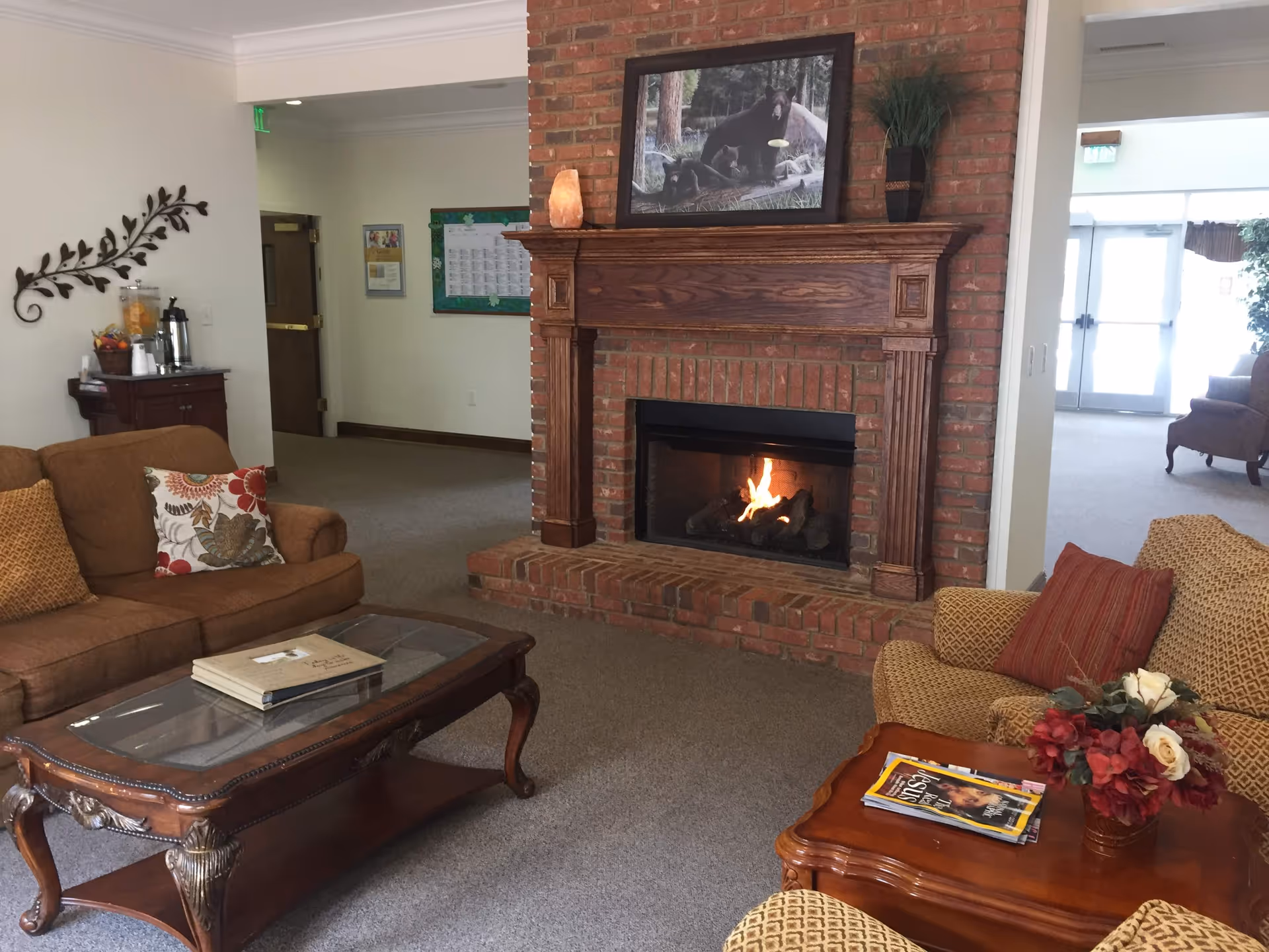 A cozy living room area with a brick fireplace featuring a wooden mantle and a fire burning inside. Above the fireplace is a framed picture of bears. The room has two upholstered sofas with decorative pillows, a wooden coffee table with a glass top, and a side table with a floral arrangement and magazines. In the background, there is a small refreshment station with a beverage dispenser and coffee pot, and a hallway leading to other rooms.