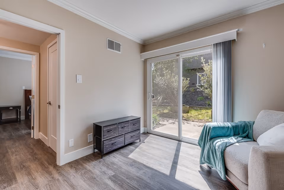 A bright living room area with a sliding glass door leading to an outdoor garden. The room features wood flooring, a beige wall, a small dark wooden cabinet with drawers, and a beige armchair with a teal throw blanket draped over it. Adjacent to the living room is a doorway leading to a bedroom with a dark wooden nightstand.