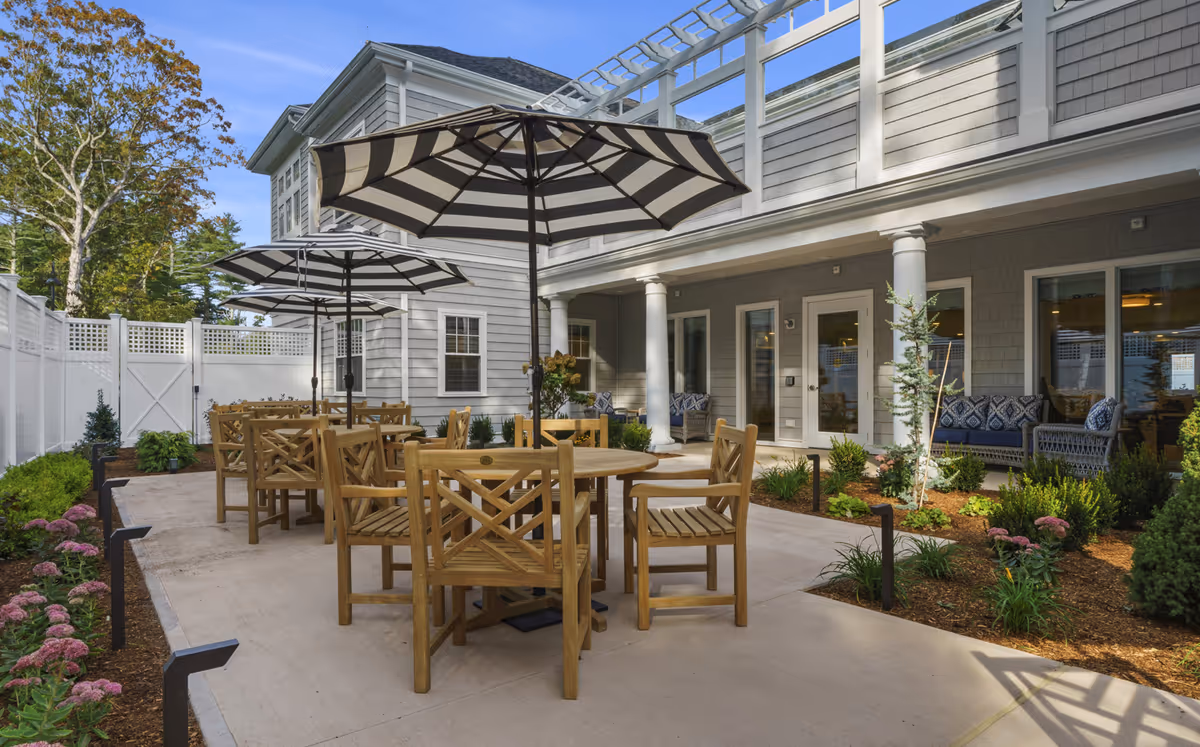 Outdoor patio area at The Village at Duxbury featuring wooden tables and chairs with large striped umbrellas. The patio is surrounded by landscaped garden beds with flowers and shrubs, and the building exterior has gray siding with white trim and columns. There are cushioned seating areas near the building under a covered porch.