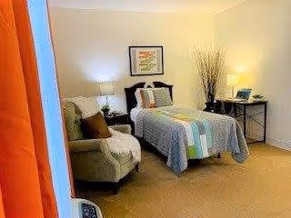 A cozy bedroom featuring a single bed with a colorful quilt, a green armchair with a pillow and blanket, a side table with a lamp, a framed picture on the wall, and a desk with a lamp and decorative tall grass in a vase.