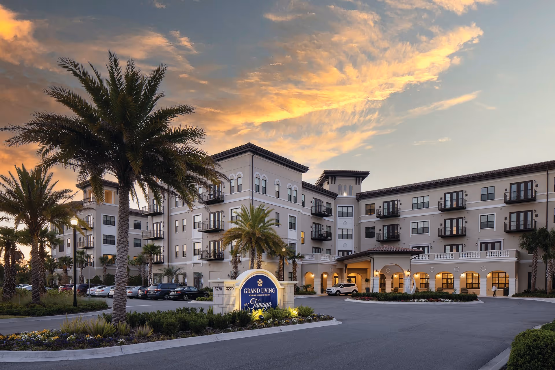 Exterior view of the Grand Living At Tamaya senior living facility at sunset, featuring a multi-story building with balconies, palm trees, a parking lot with cars, and a prominent entrance sign illuminated by lights.