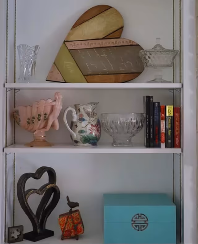 A white shelving unit displaying decorative items including a large heart-shaped wall art, glassware, a floral pitcher, a pink ceramic bowl with a figure, a set of books, a black heart-shaped sculpture, a small bird figurine, a colorful pouch, and a turquoise box with an Asian-inspired symbol.