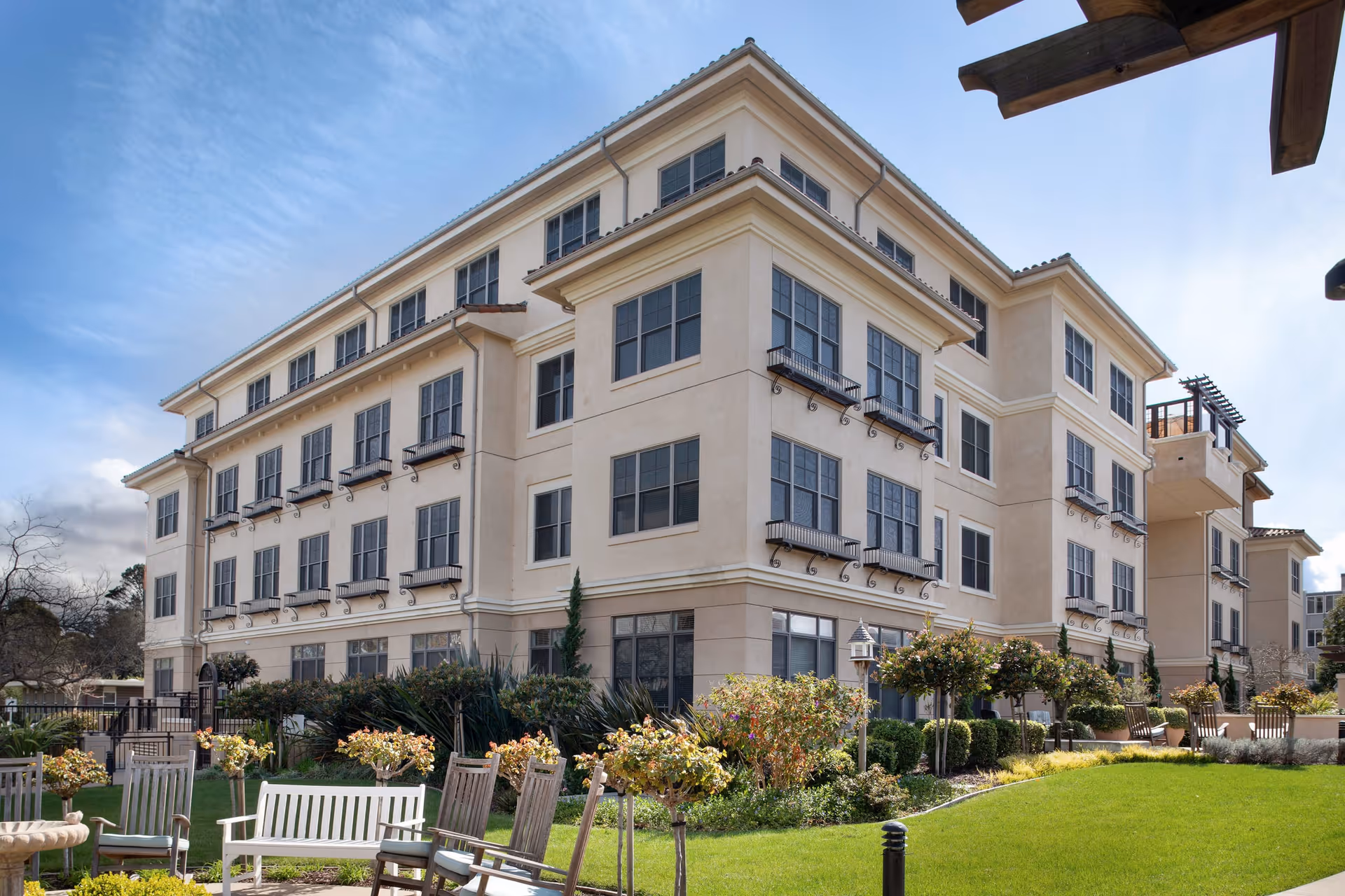 Exterior view of a multi-story senior living facility building with large windows and beige walls. In the foreground, there is a well-maintained garden with green grass, shrubs, small trees, and several wooden chairs arranged in a seating area under a clear blue sky.