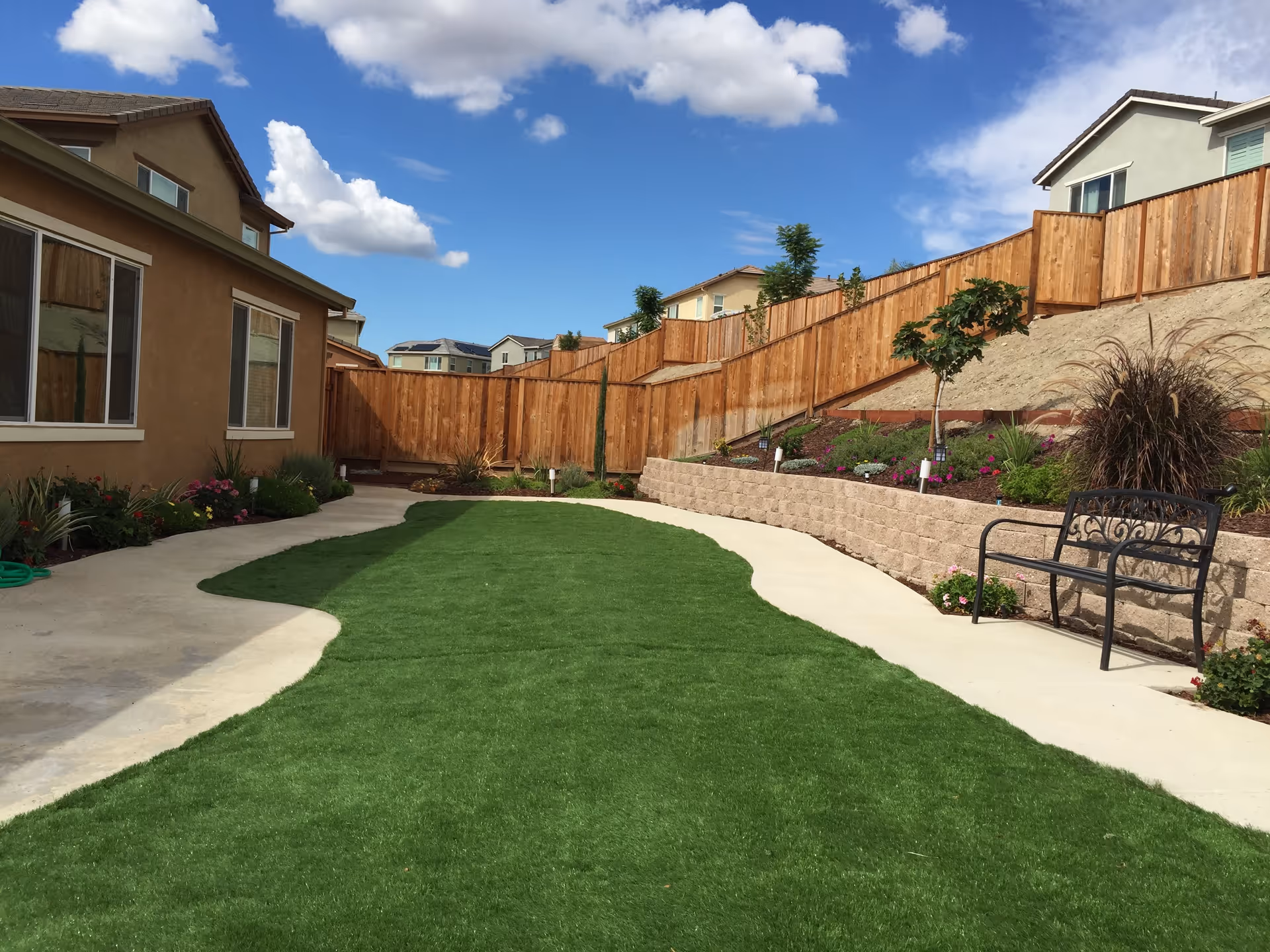 Outdoor garden area with a green lawn, concrete pathways, a wooden fence, various plants and flowers, a metal bench, and houses in the background under a partly cloudy blue sky.