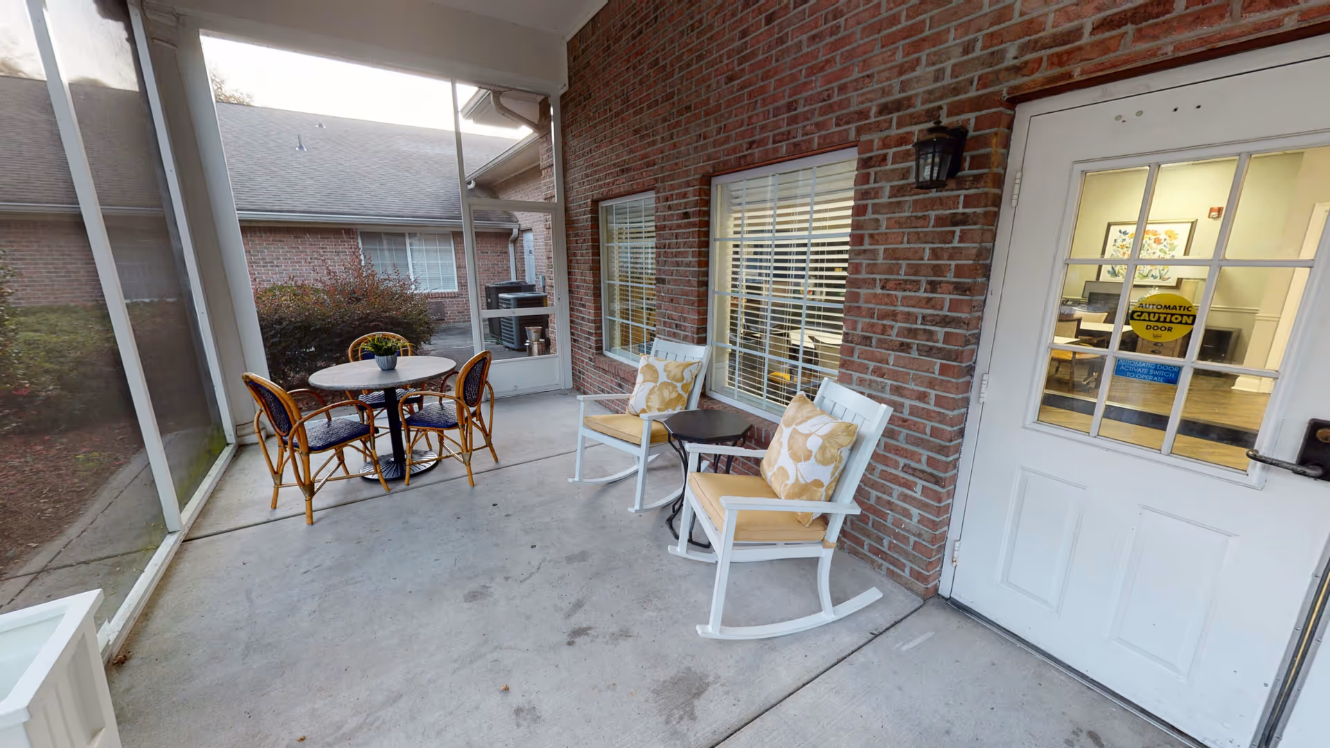 A screened-in porch area with two white rocking chairs with yellow cushions and floral pillows, a small black side table between them, and a round table with four wicker chairs with blue cushions. The porch has a concrete floor and brick walls with two windows. A white door with glass panes leads inside, showing an interior room with tables, chairs, and a framed floral artwork on the wall.
