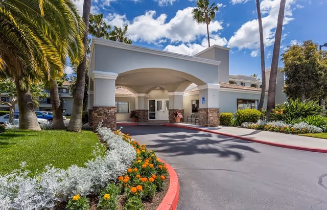 Entrance of a senior living facility named Las Villas Del Norte with a covered drop-off area, surrounded by palm trees, green grass, and colorful flowers under a partly cloudy blue sky.