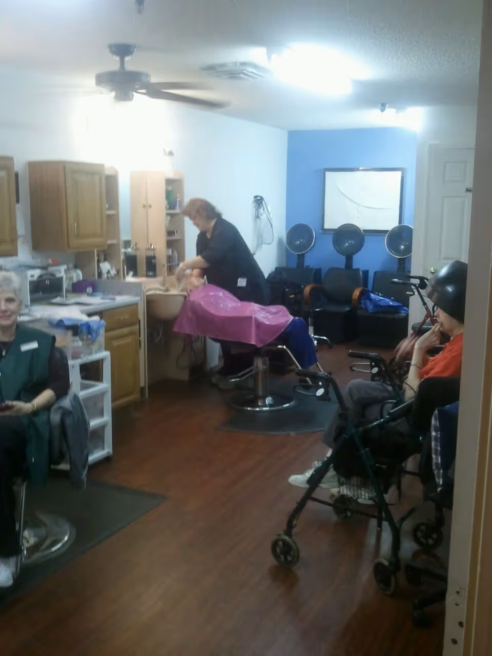 Interior of a salon area in a senior living facility with a hairdresser washing an elderly person's hair at a sink. Another elderly person is sitting under a hair dryer, and a third person is seated nearby. The room has wooden floors, cabinets, and salon chairs.