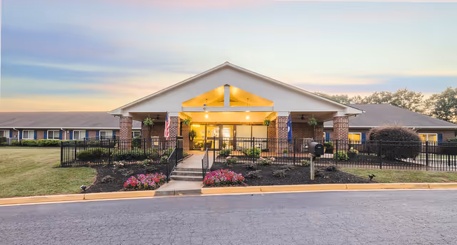 Front entrance of a single-story brick post-acute facility with a covered entryway, lit interior, landscaped flower beds, and flags.