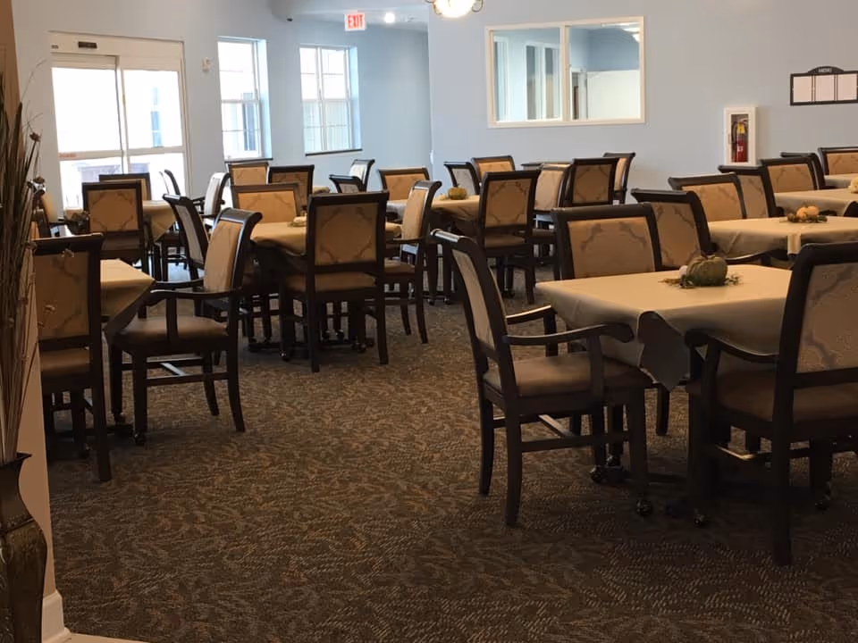 A dining room in Symphony Assisted Living and Memory Support with multiple tables covered with beige tablecloths and surrounded by cushioned chairs. The room has carpeted flooring, light blue walls, and windows letting in natural light. There are small decorative centerpieces on the tables.