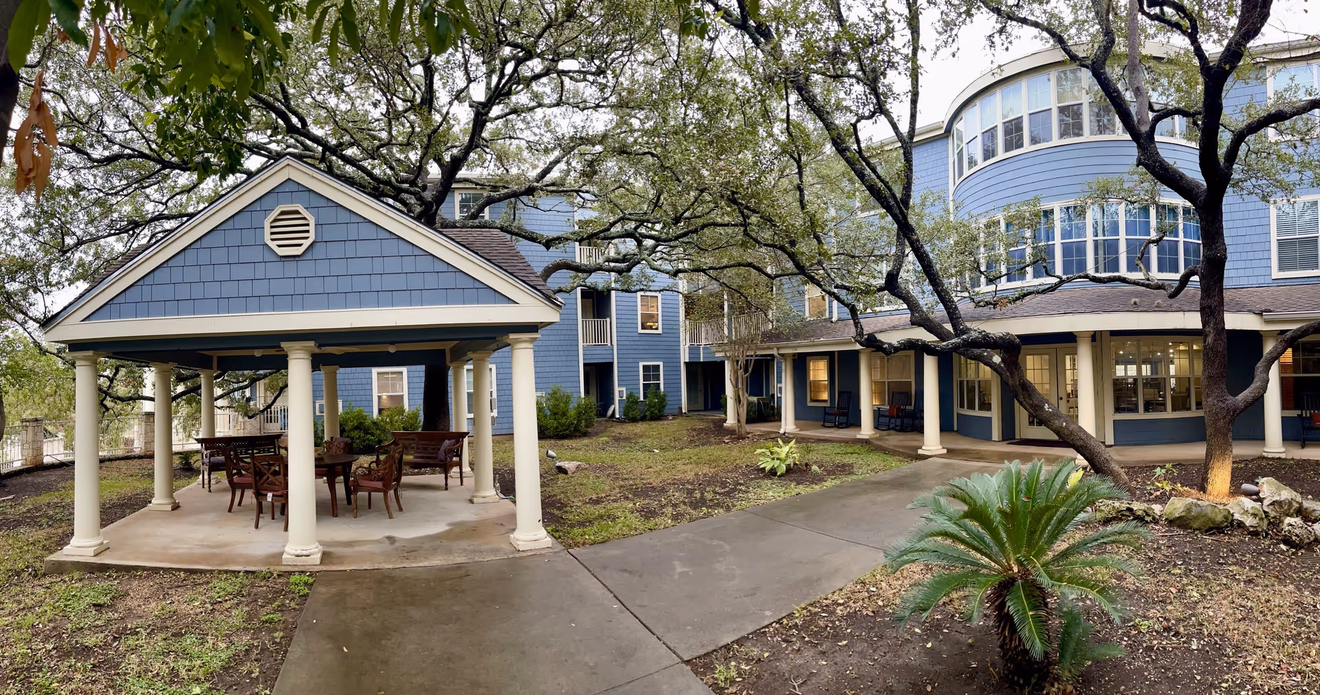 Outdoor area of Independence Hill Retirement Community featuring a covered pavilion with tables and chairs, surrounded by trees and landscaping. The blue building with white trim and multiple windows is visible in the background.