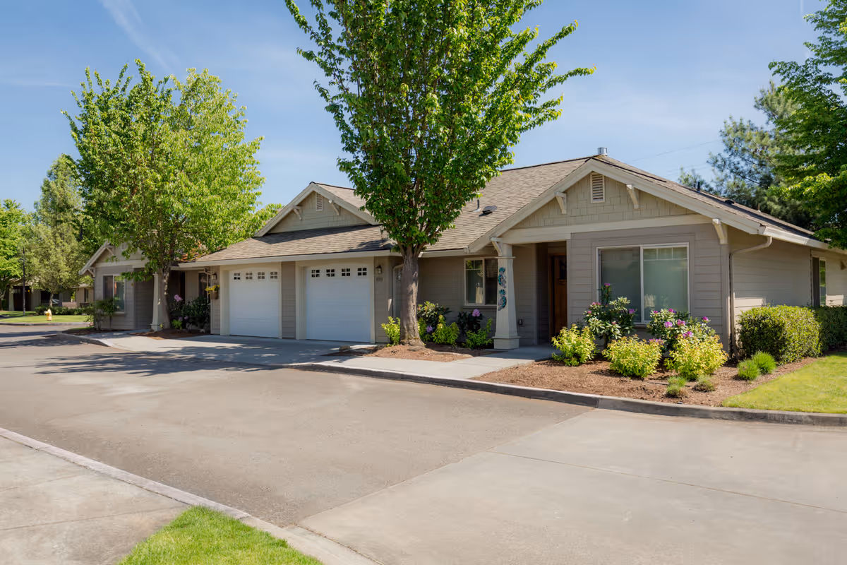 Exterior view of a single-story residential building with beige siding, two white garage doors, and a small front porch. The building is surrounded by green trees, shrubs, and a well-maintained lawn under a clear blue sky.