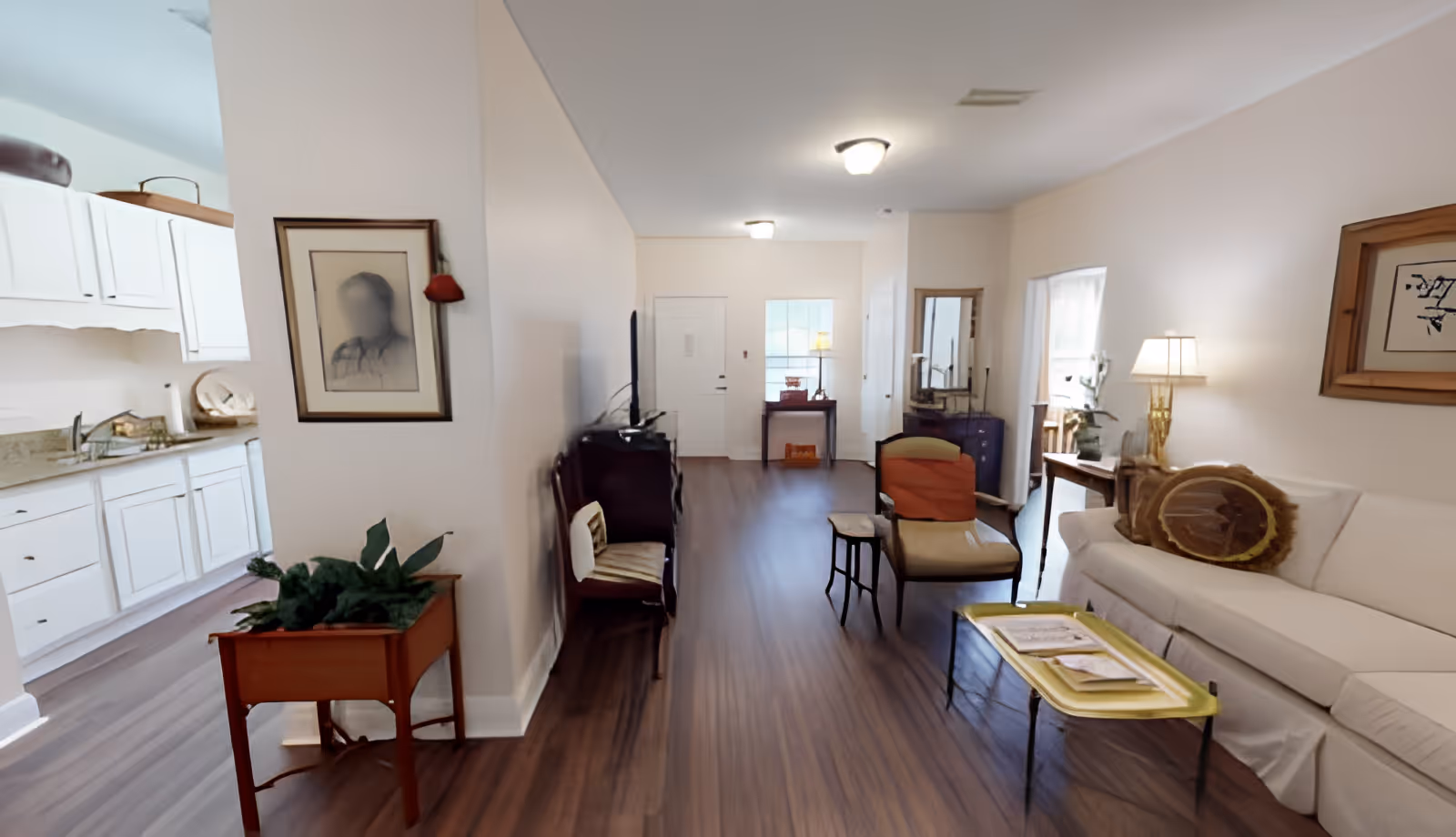 Interior view of a senior living facility showing a living room with a white sofa, an armchair, a small coffee table with magazines, and a lamp. The room has wooden flooring and light-colored walls. To the left, there is a partial view of a kitchen with white cabinets and a countertop. The entrance door and a small table with a lamp are visible in the background.