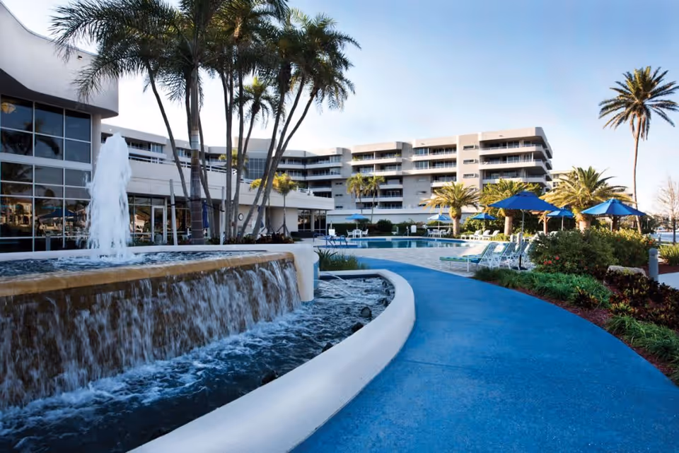 Outdoor view of Harbour's Edge facility featuring a cascading water fountain, palm trees, a blue walking path, lounge chairs with blue umbrellas, and a multi-story building in the background under a clear sky.