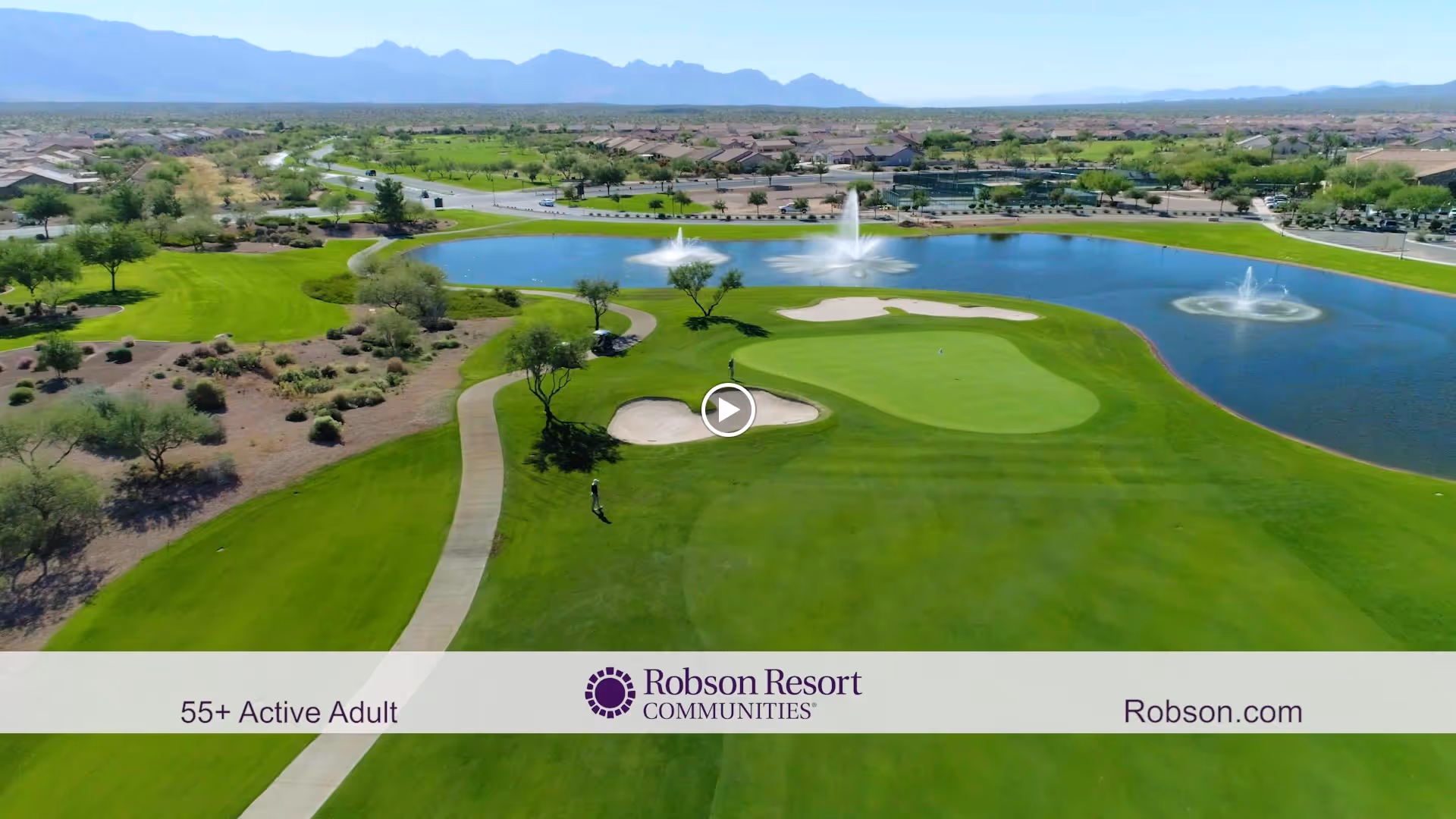 Aerial view of a golf course with green fairways, sand bunkers, and a large water feature with fountains. In the background, there are residential houses and mountains under a clear sky.