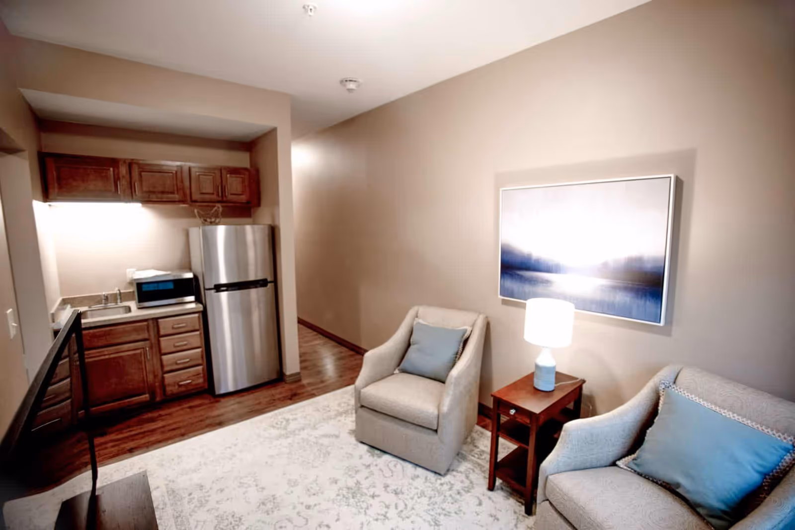 A small living area with two beige armchairs, each with a blue cushion, a wooden side table with a white lamp, and a framed abstract painting on the wall. To the left, there is a compact kitchen area with wooden cabinets, a stainless steel refrigerator, a microwave, and a sink. The floor is wooden with a light-colored patterned rug in the living area.