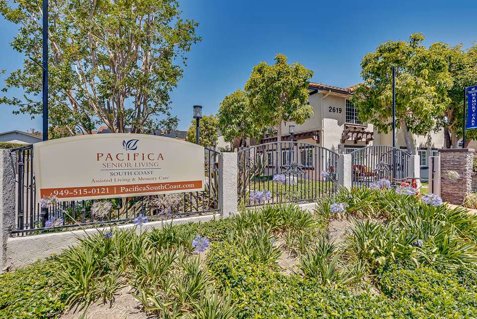 Exterior view of Pacifica Senior Living South Coast facility with a sign displaying the name, phone number, and website. The area is landscaped with green plants and purple flowers, and there are trees and a gated entrance visible under a clear blue sky.